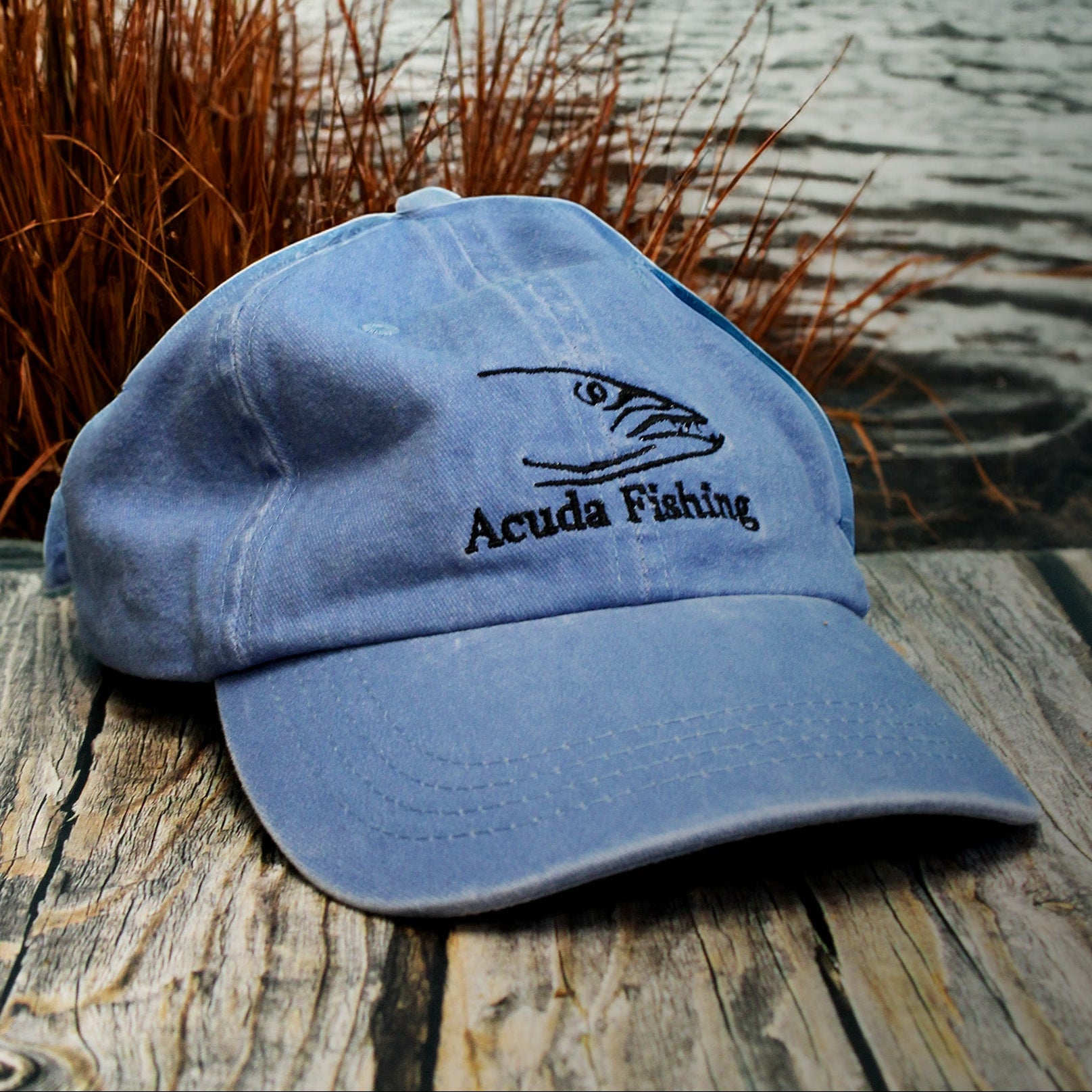 Blue cap with 'Acuda Fishing' logo on a wooden surface near a lake with mountains in the background